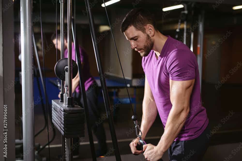 Young adult bodybuilder healthy fitness man doing triceps overhead extension rope exercise. Workout in public gym and sporty lifestyle concept